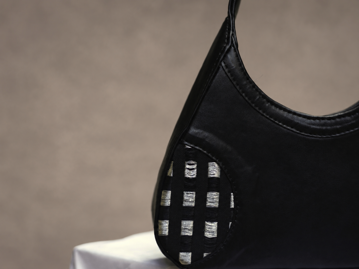 Black Orisun leather handbag with a woven black-and-silver panel, on white cloth against a soft taupe backdrop.