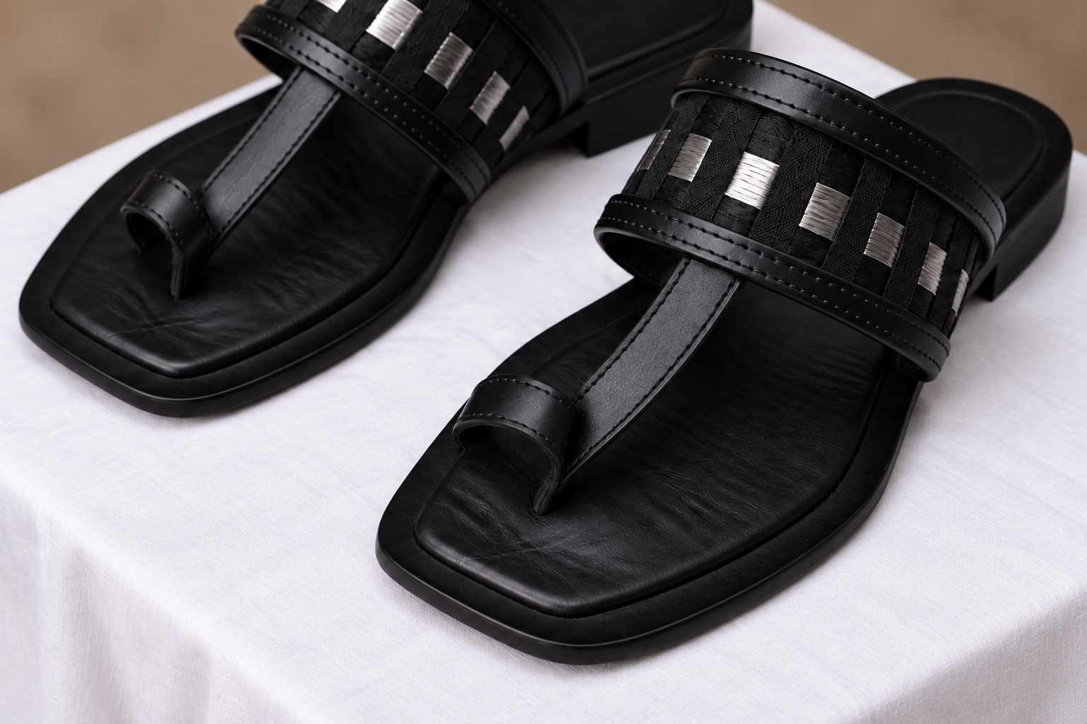 A pair of black leather Orisun toe-ring slippers with woven black-and-silver straps, on white cloth.